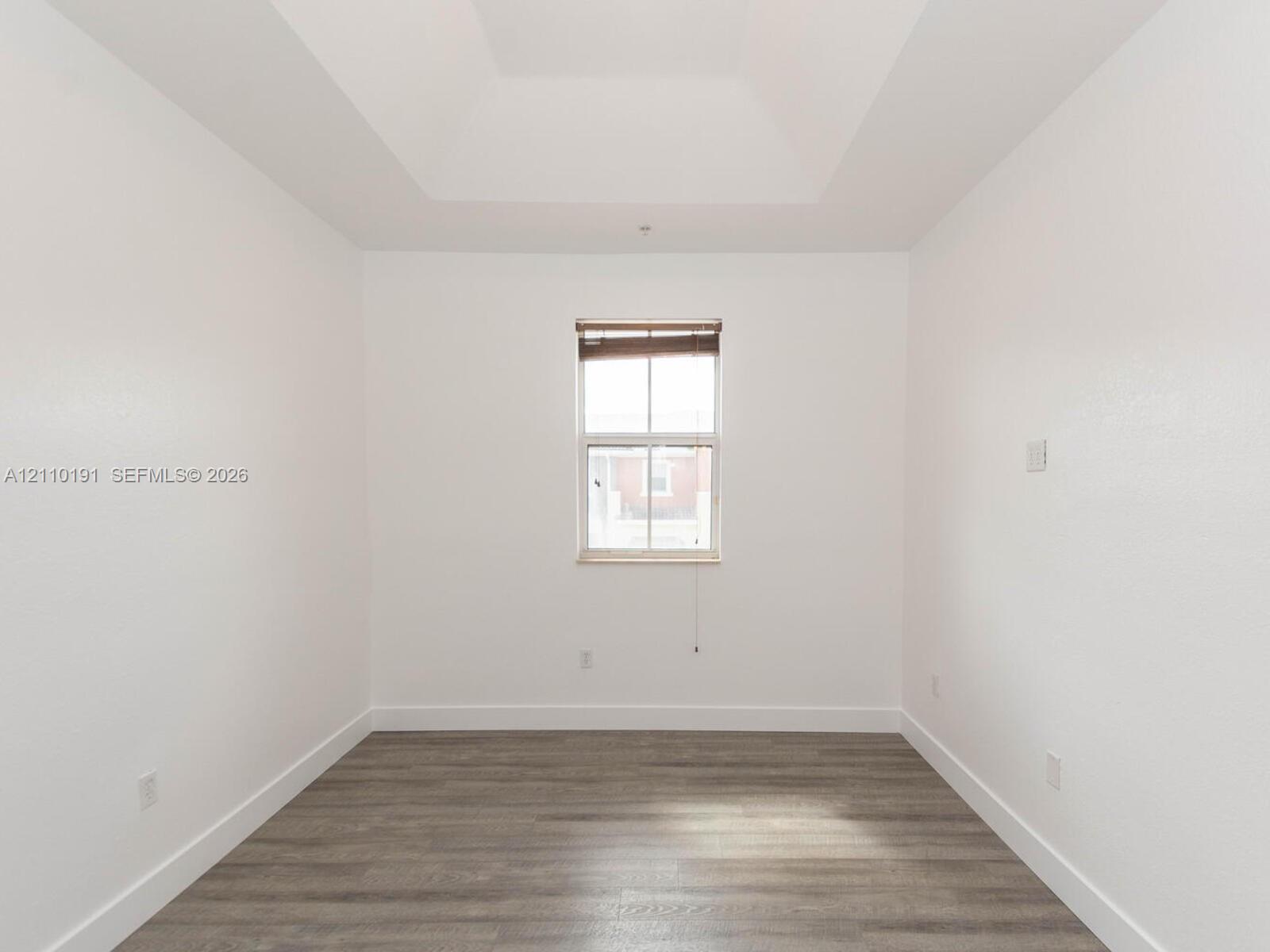 Room featuring tray ceilings, wood-finish flooring, and a central window with integrated blinds