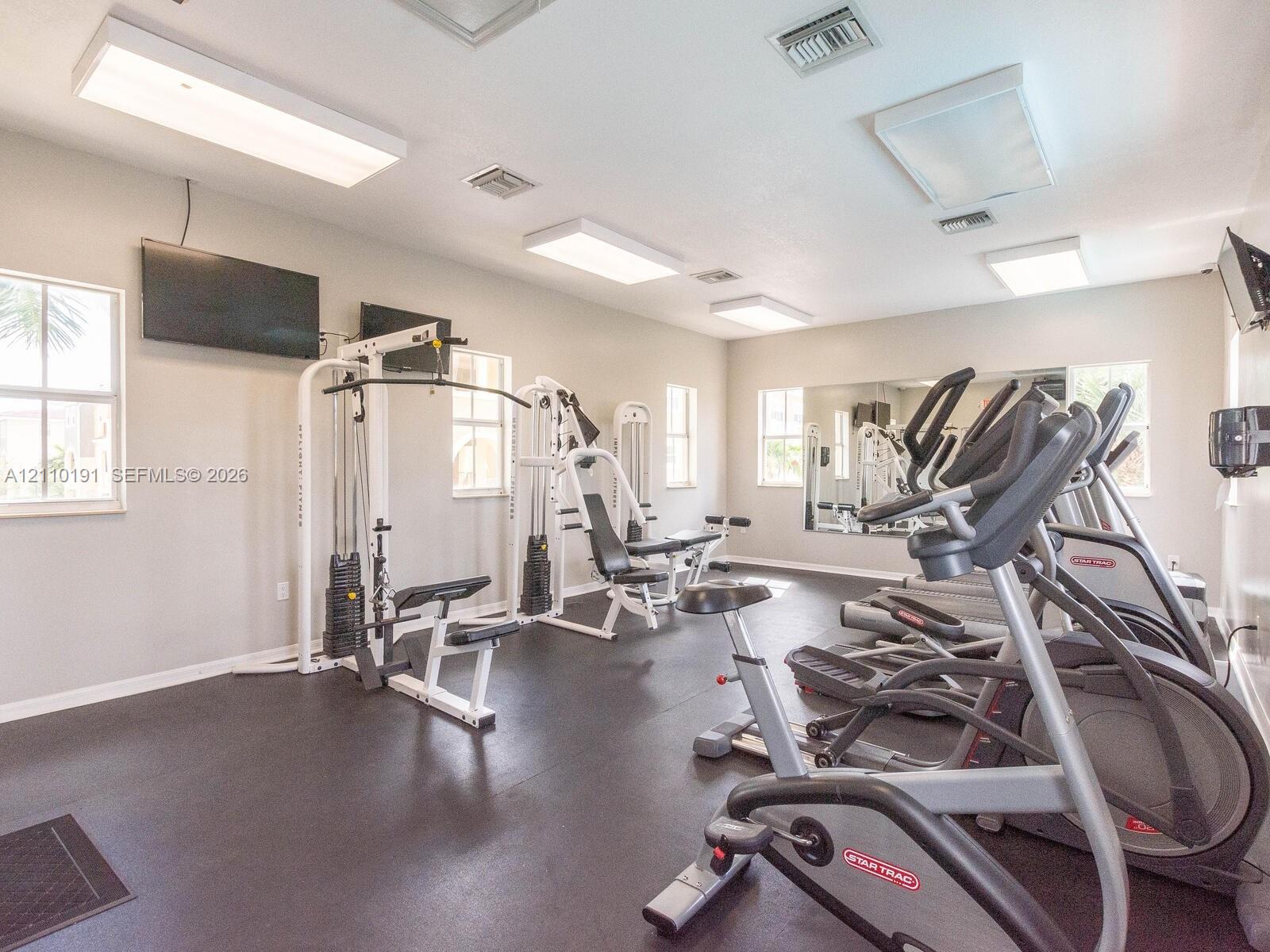 Fitness center featuring rubberized flooring, large wall-mounted mirrors, multiple overhead panel lights, and windows