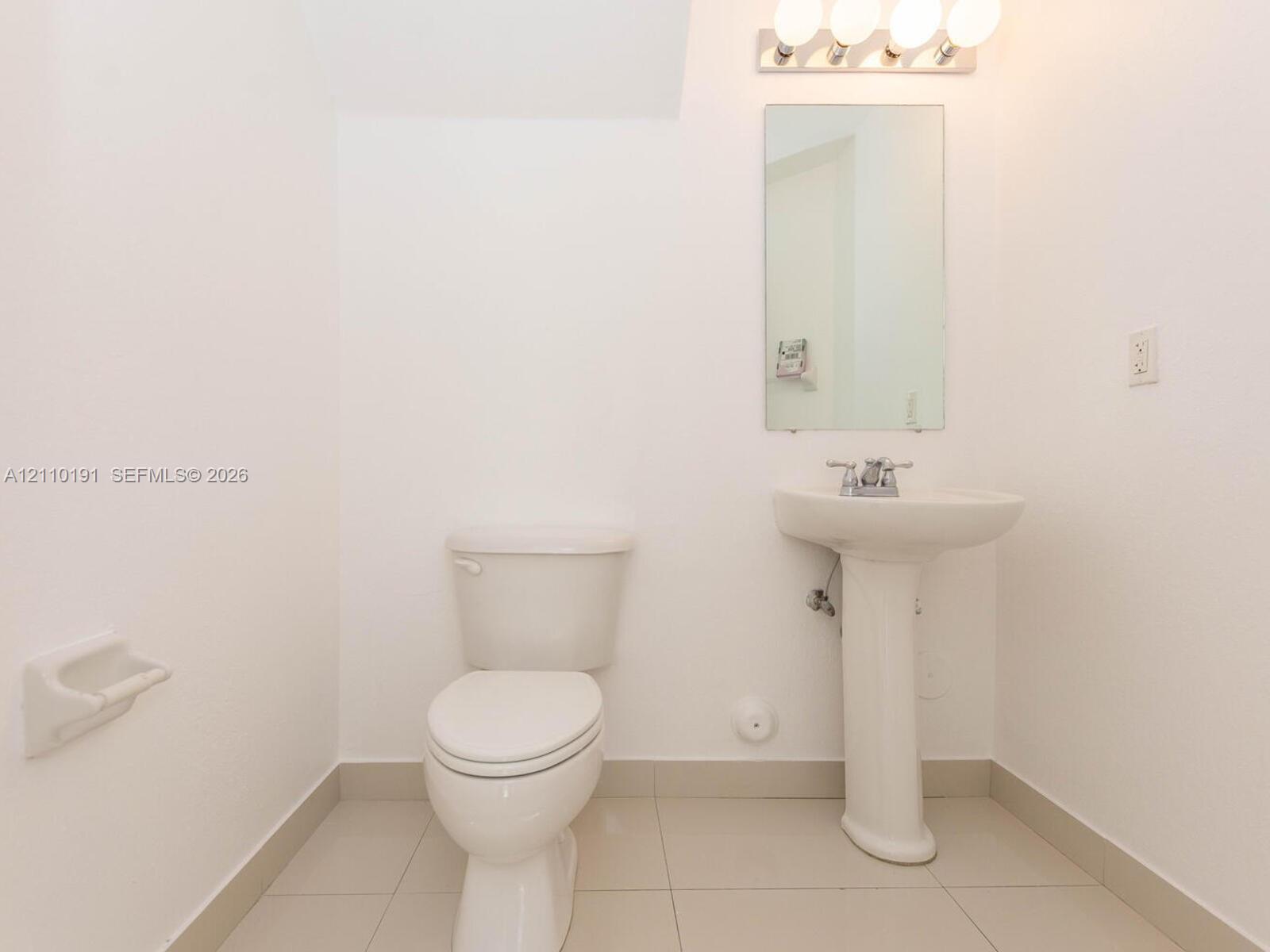Bathroom featuring a white pedestal sink, dual-handle faucet, wall-mounted rectangular mirror, four-light vanity fixture, and tiled flooring