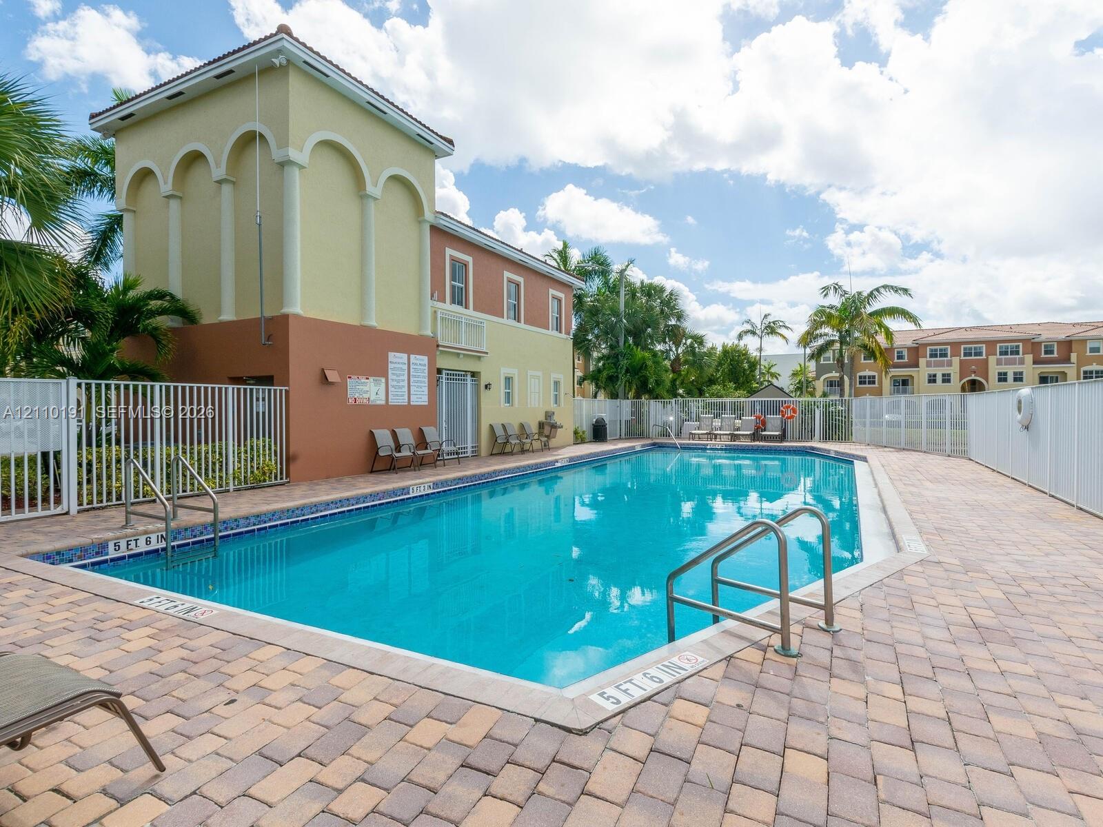 Swimming pool with tiled waterline, concrete paver decking, metal perimeter fencing, and two-story community building