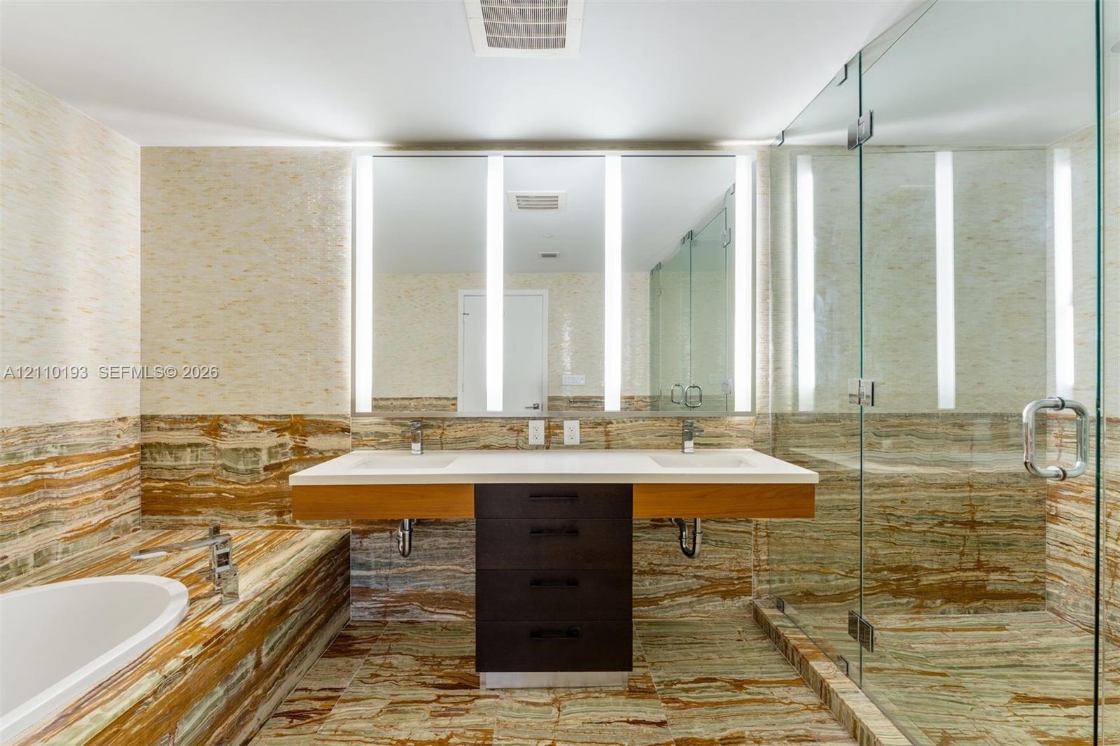 Floating double vanity with a stone countertop, integrated LED-lit mirror, soaking tub, and a glass-enclosed walk-in shower featuring extensive stone wall and floor tiling