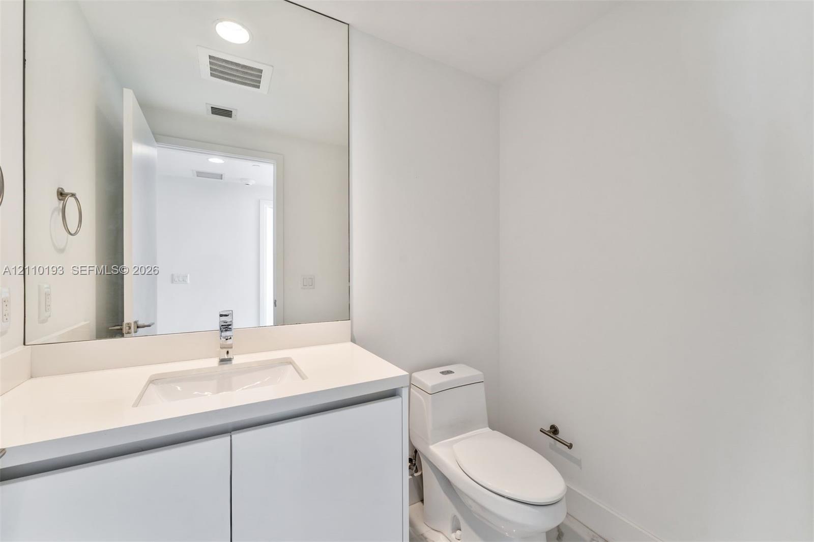 White vanity with solid-surface countertop, rectangular integrated sink, chrome single-handle faucet, full-width mirror, and dual-flush toilet