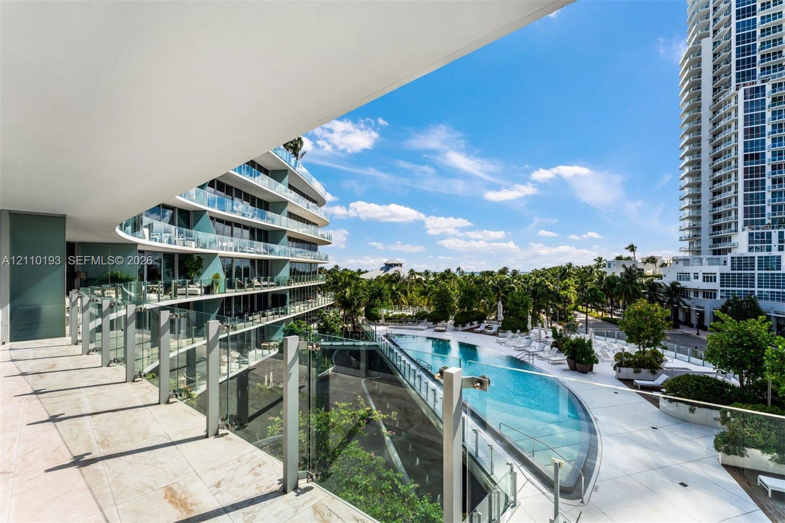 Covered terrace featuring stone tile flooring and glass panel railings, overlooking a swimming pool with a stone deck, mature palm trees, and structural landscape elements Covered terrace featuring stone tile flooring and glass panel railings, overlooking a swimming pool with a stone deck, mature palm trees, and structural landscape elements