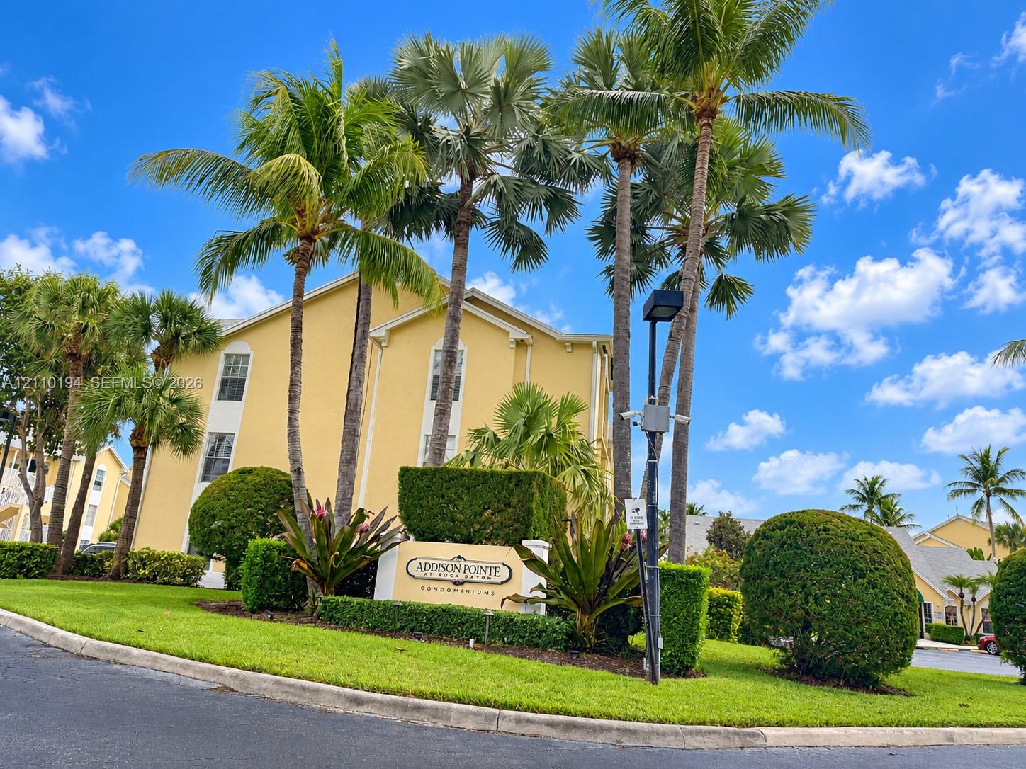 Multi-story condominium exterior featuring stucco siding, mature palm trees, and professional landscaping with manicured shrubbery