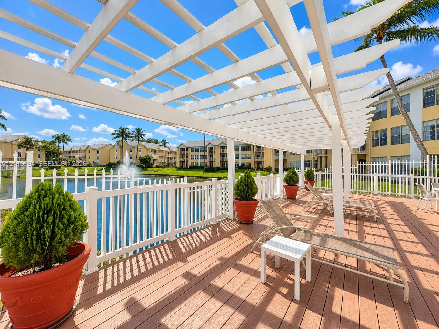 Expansive wood-tone deck featuring a white overhead pergola and white picket-style perimeter fencing overlooking a pond with a built-in fountain