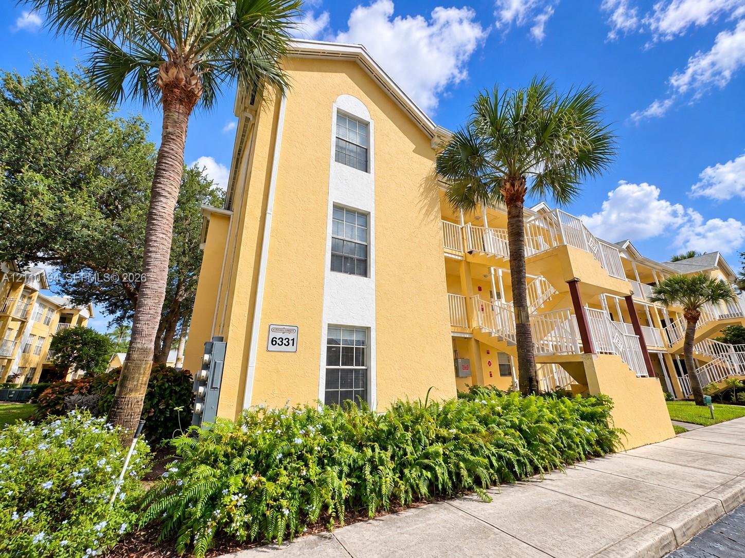 Multi-story exterior featuring a yellow stucco facade, white trim accents, arched window detailing, tiered exterior staircases with white railings, and mature palm tree landscaping Multi-story exterior featuring a yellow stucco facade, white trim accents, arched window detailing, tiered exterior staircases with white railings, and mature palm tree landscaping