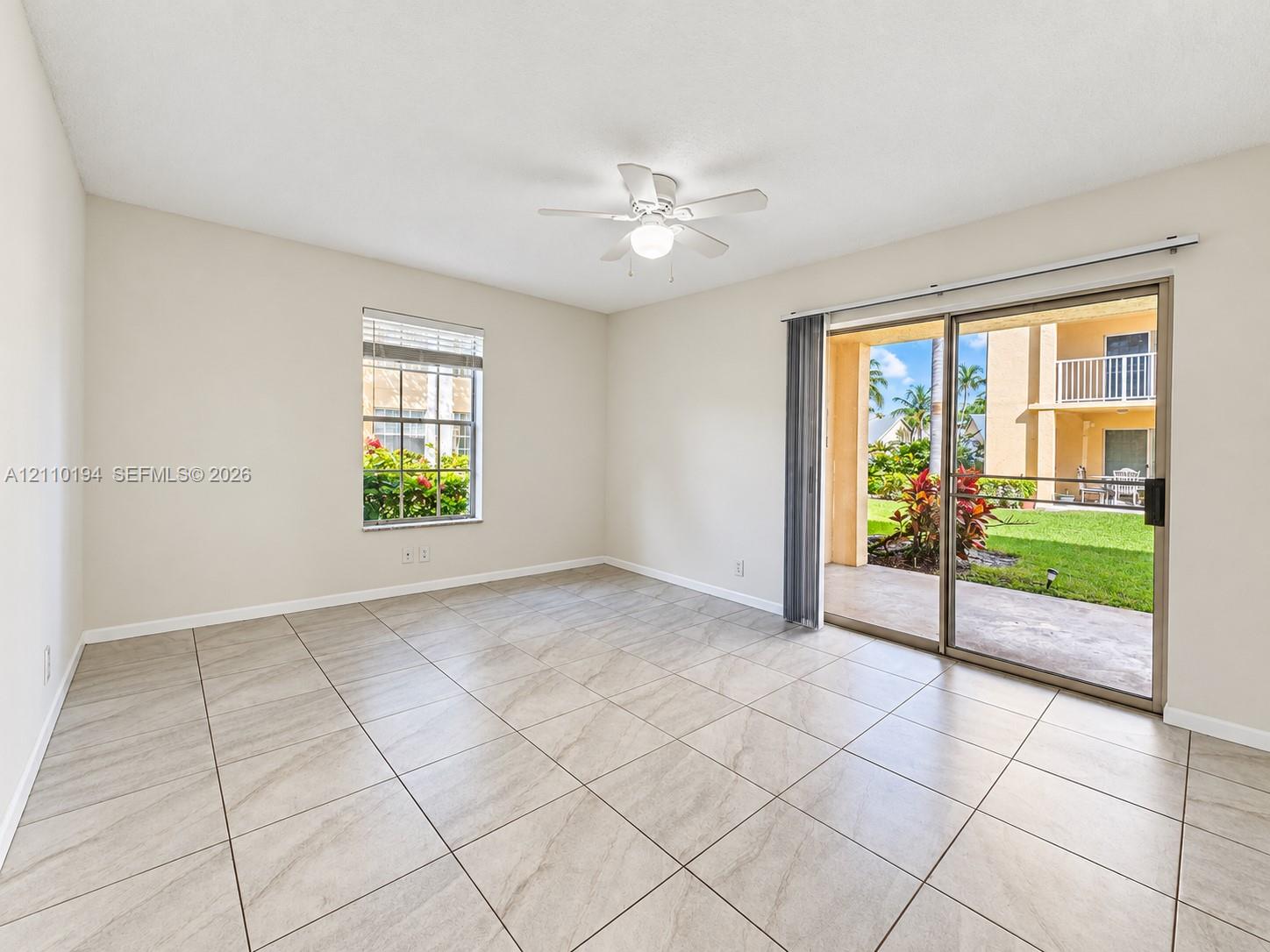 Neutral-toned interior space featuring large-format ceramic tile flooring, a ceiling fan with integrated lighting, and glass sliding doors providing access to a paved patio and outdoor space
