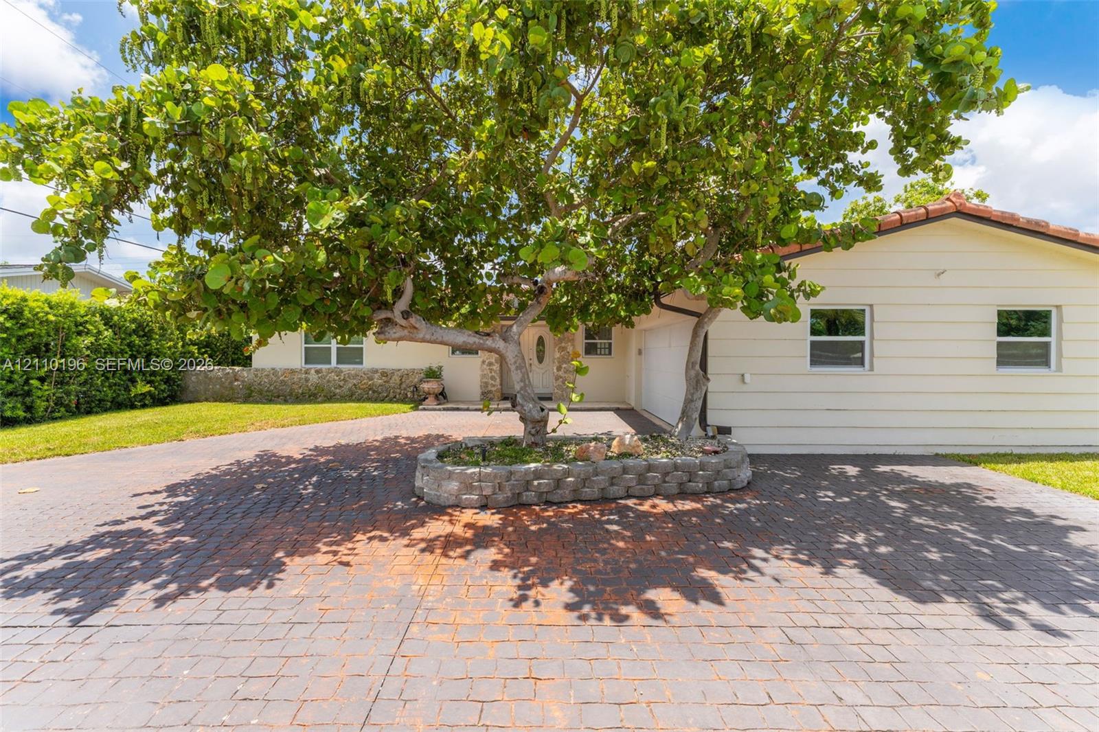 Exterior featuring a multi-tone paved driveway, a centered raised stone planter with a mature tree, horizontal siding, and a tile roof