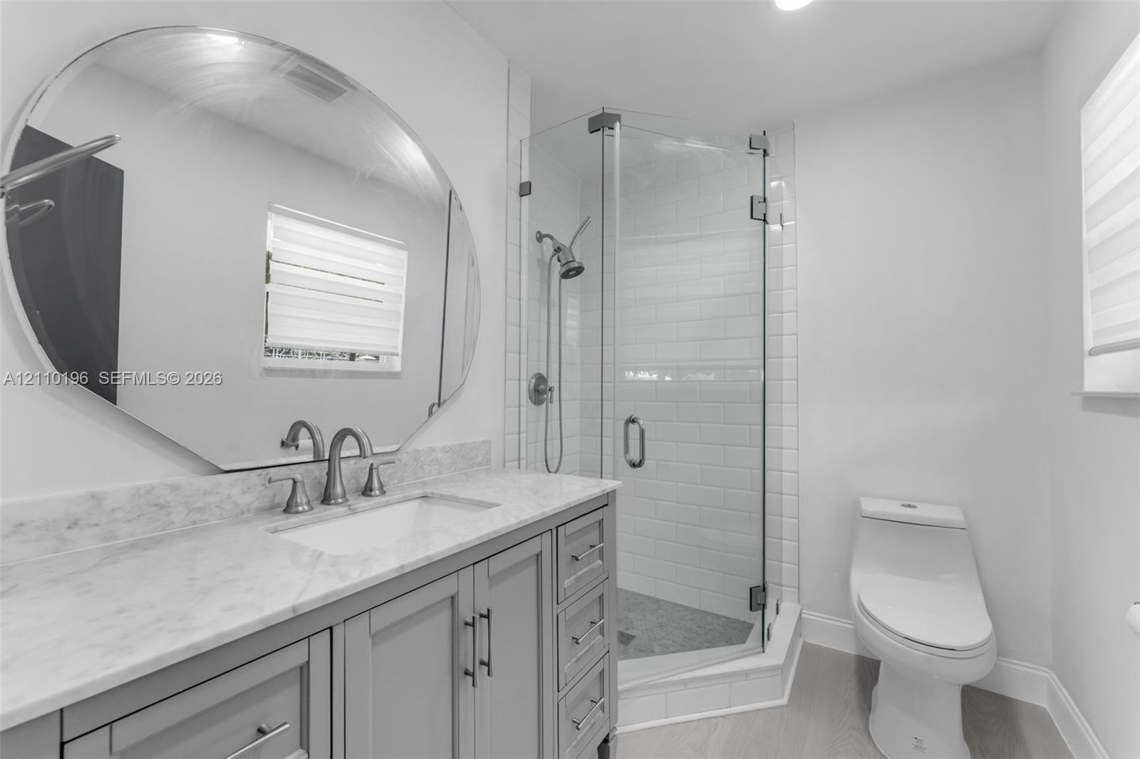 Bathroom featuring a gray vanity with a neutral stone countertop, an oversized circular mirror, a glass-enclosed shower with subway tile surround, and wood-finish flooring