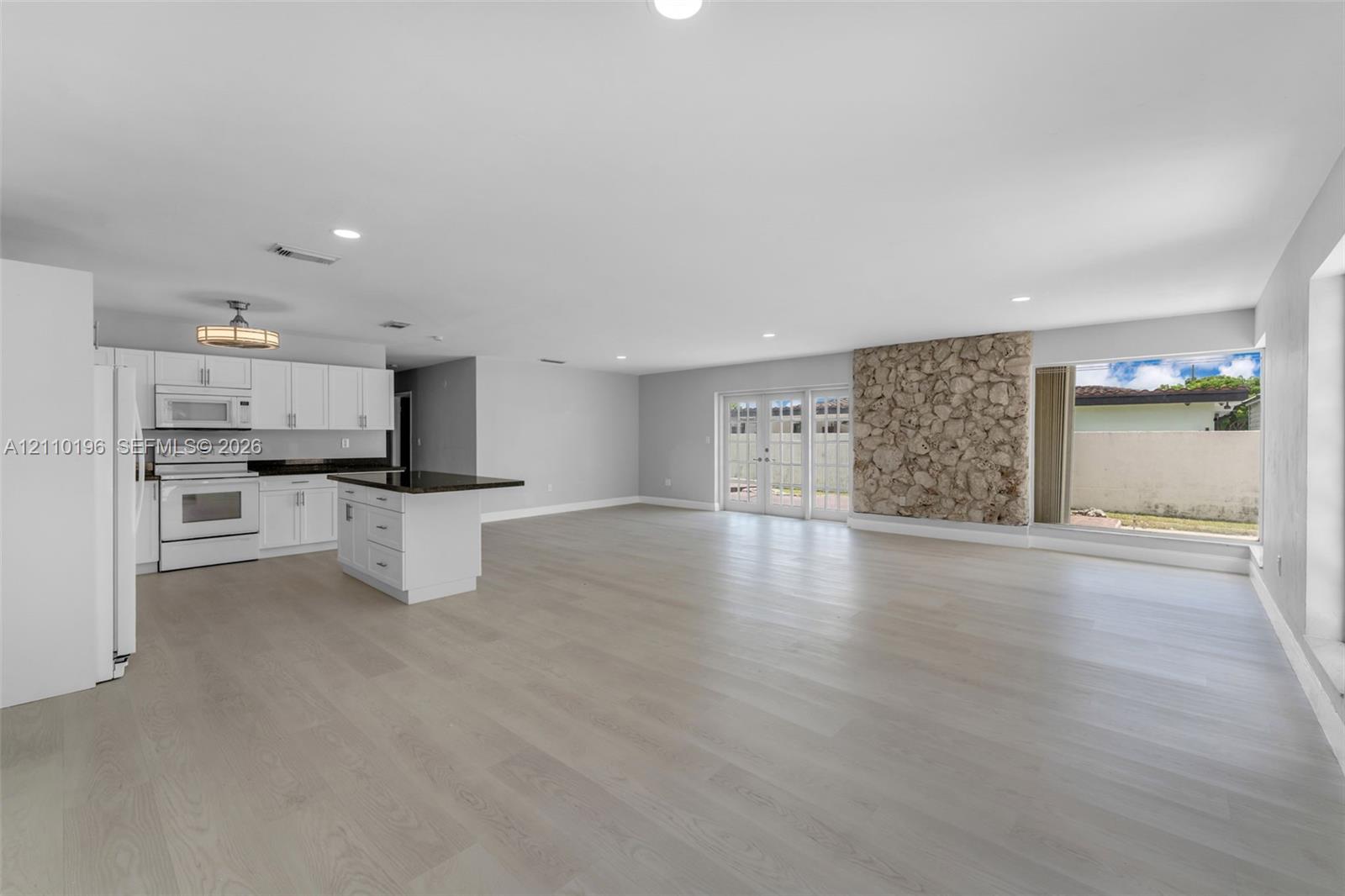 Open-concept interior featuring wood-finish flooring, a kitchen with white cabinetry and dark countertops, a natural stone accent wall, recessed lighting, and glass-paneled double doors