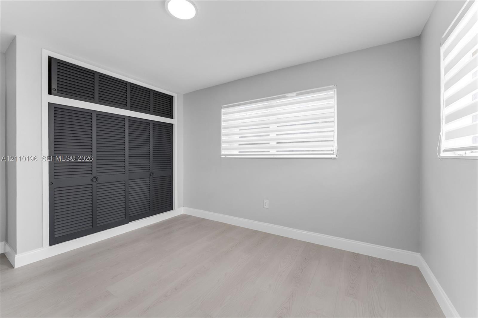 Neutral-toned room featuring wood-finish flooring, built-in floor-to-ceiling louvered-style closet cabinetry with overhead storage, and dual windows with fitted horizontal blinds