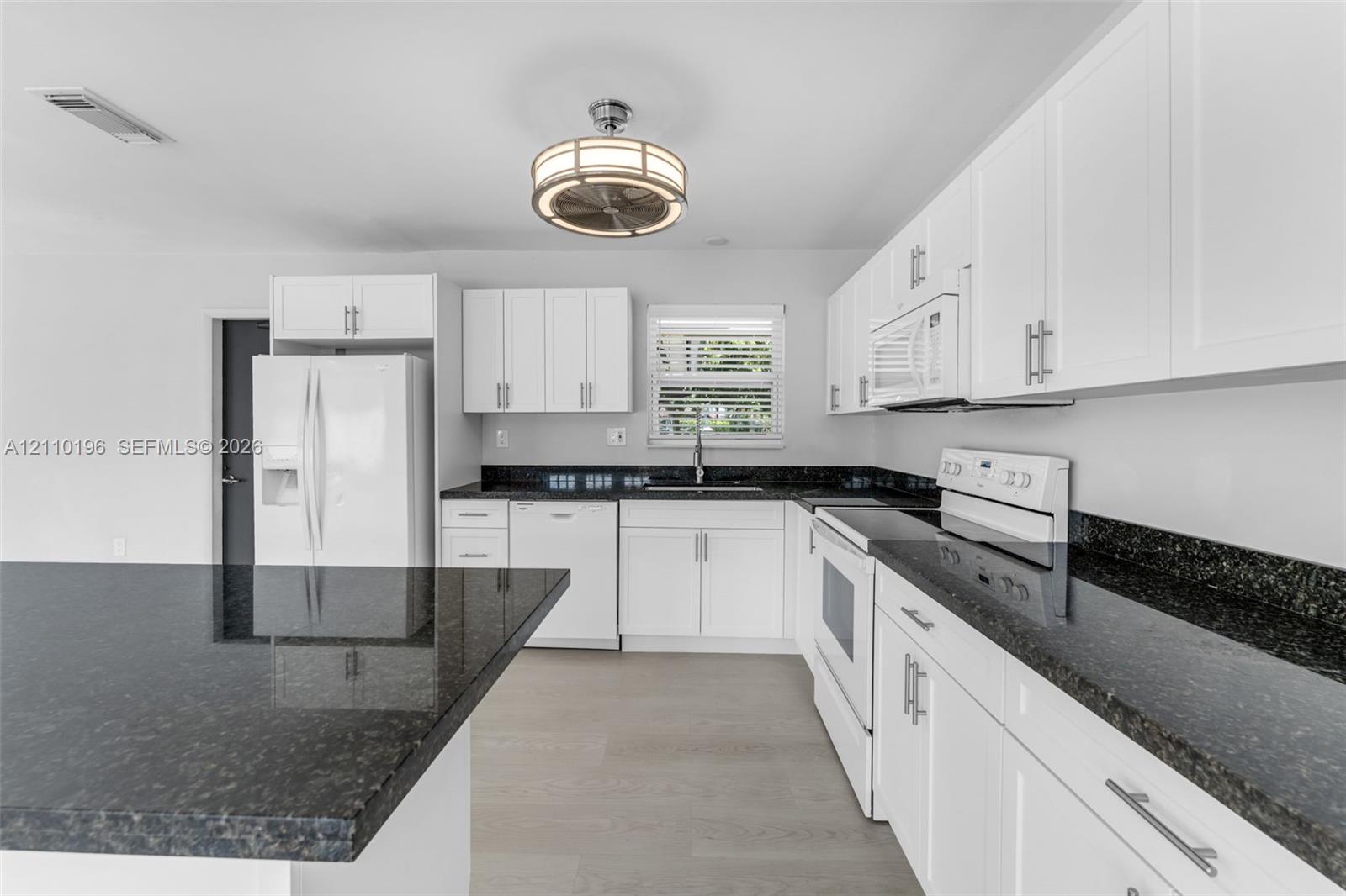 Kitchen featuring white shaker-style cabinetry, dark stone countertops, an integrated island, wood-finish flooring, and a drum-style ceiling light fixture
