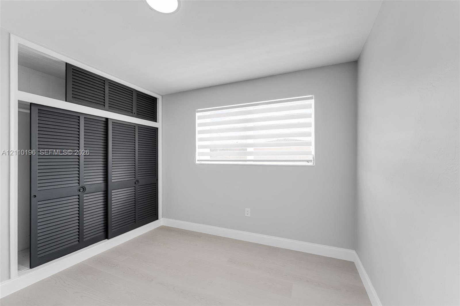 Interior space featuring light-toned wood-finish flooring, charcoal-colored louvered sliding closet doors with built-in overhead storage, and dual-layered adjustable window blinds