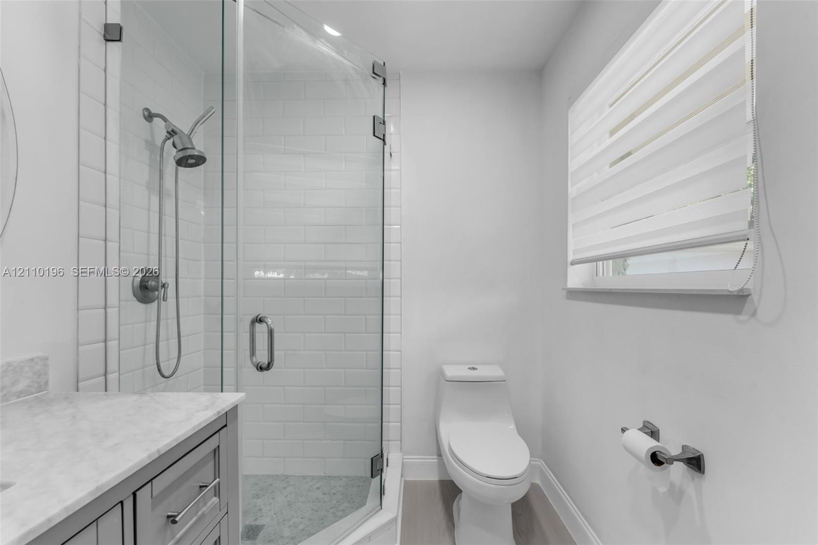 Walk-in shower featuring white subway tile surround, glass enclosure, and hand-held shower fixture, paired with a stone-top vanity and one-piece toilet
