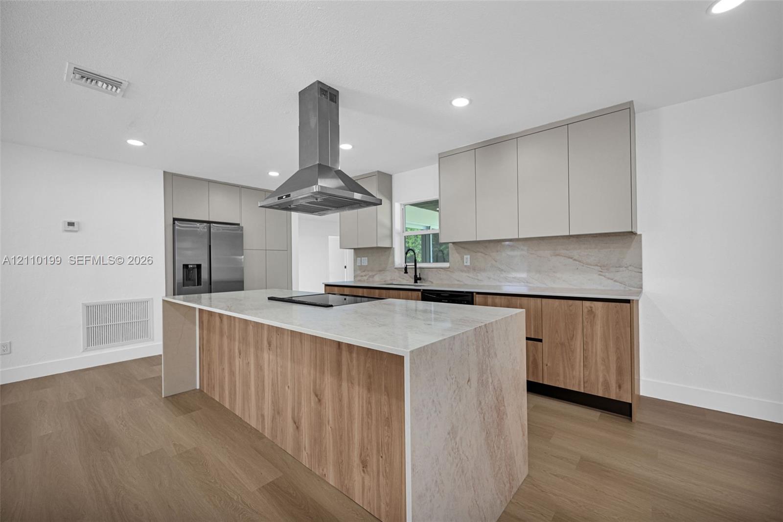 Kitchen featuring flat-panel two-tone cabinetry, central island with waterfall countertop, stainless steel chimney-style range hood, stone-finish backsplash, and wood-finish flooring