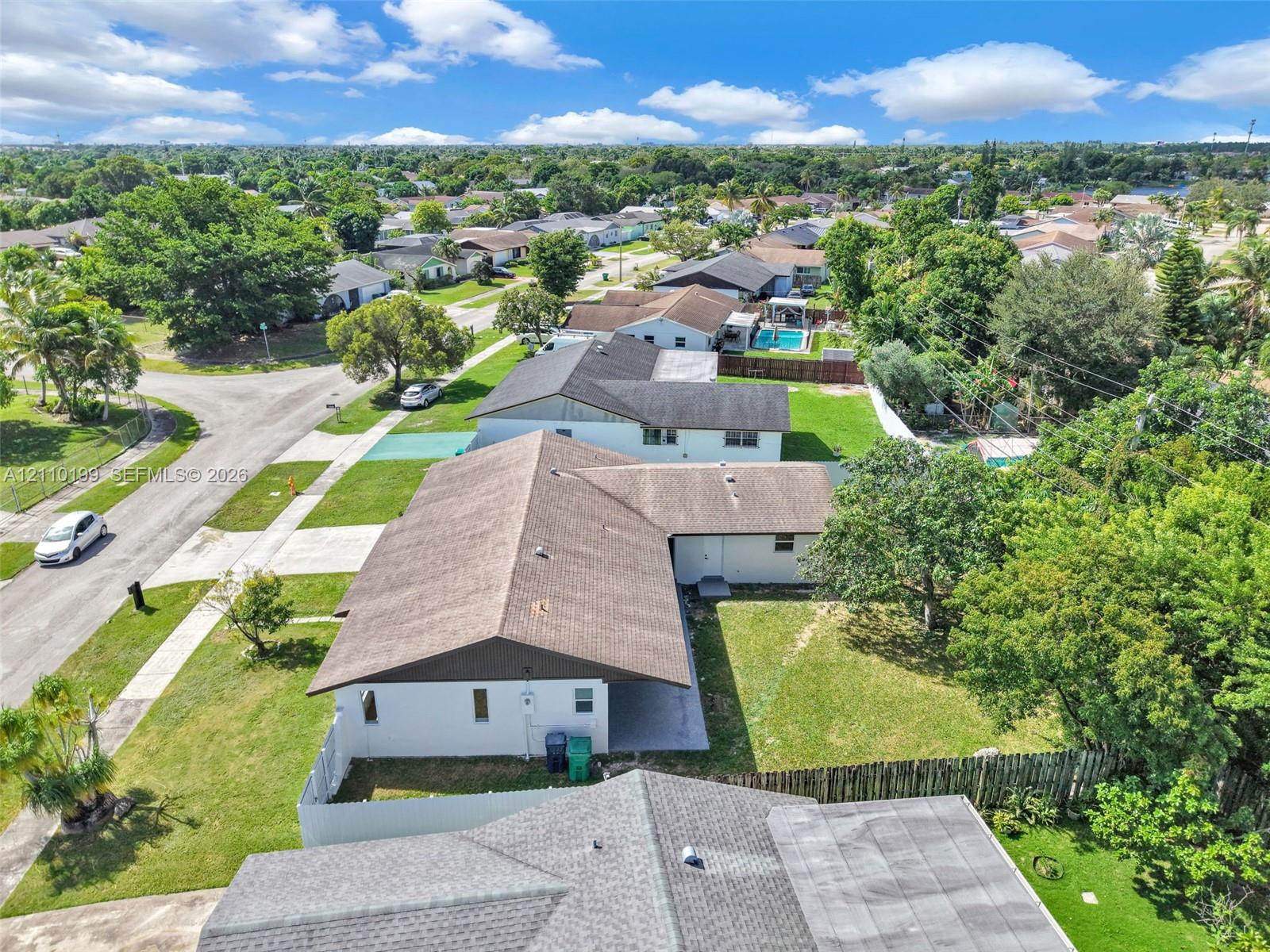 Aerial view featuring a multi-pitched shingle roof, white exterior siding, concrete driveway, and a fenced backyard with mature trees