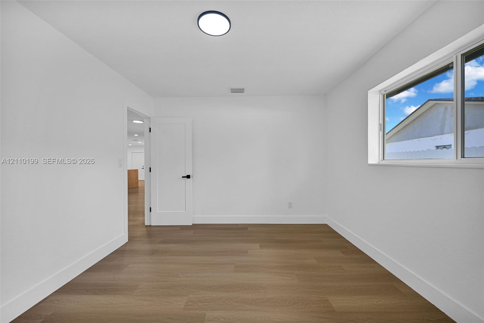 Room featuring wood-finish flooring, crisp white walls, a dual-pane sliding window, a modern flat-panel door with matte black hardware, and a flush-mount LED light fixture