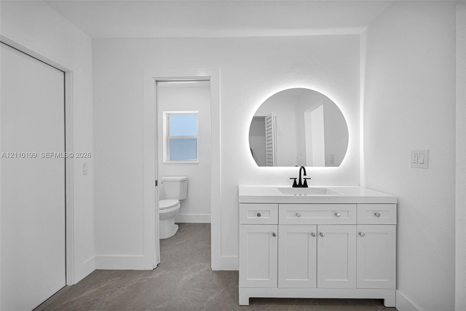 White vanity featuring a solid-surface countertop with an integrated rectangular sink and matte black faucet, illuminated semi-circular wall mirror, and neutral stone-finish tile flooring