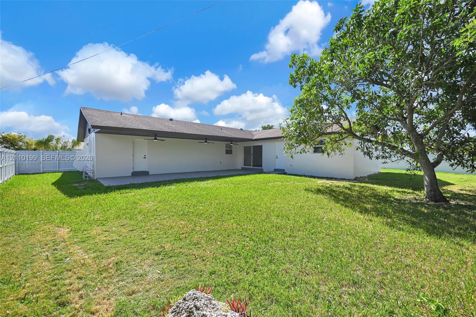 Expansive backyard featuring a manicured lawn, mature trees, a covered patio with ceiling fans, and a white privacy fence
