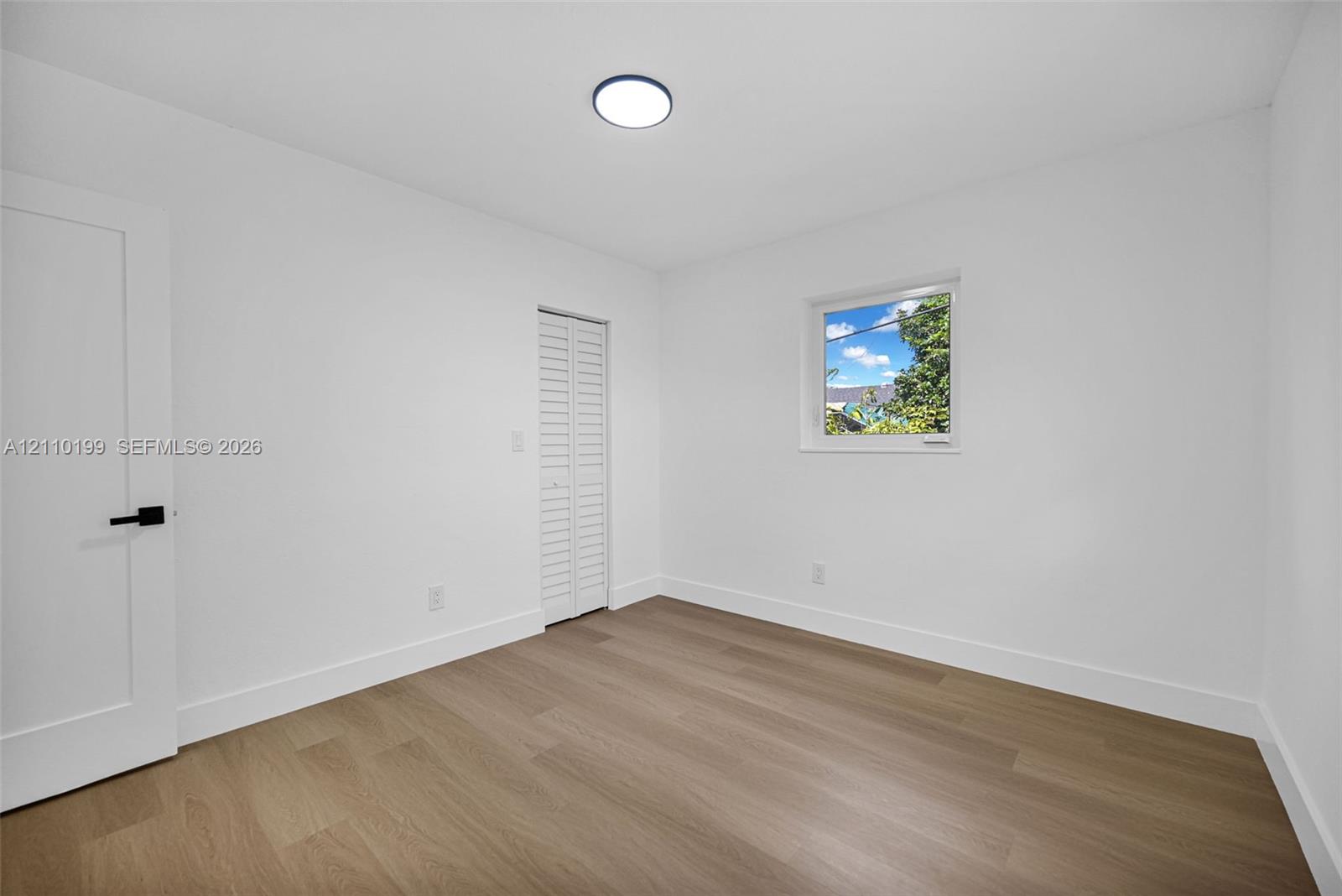 Interior space featuring wood-finish flooring, white walls, a single-window configuration, flush-mount lighting, and a closet with white louvered doors