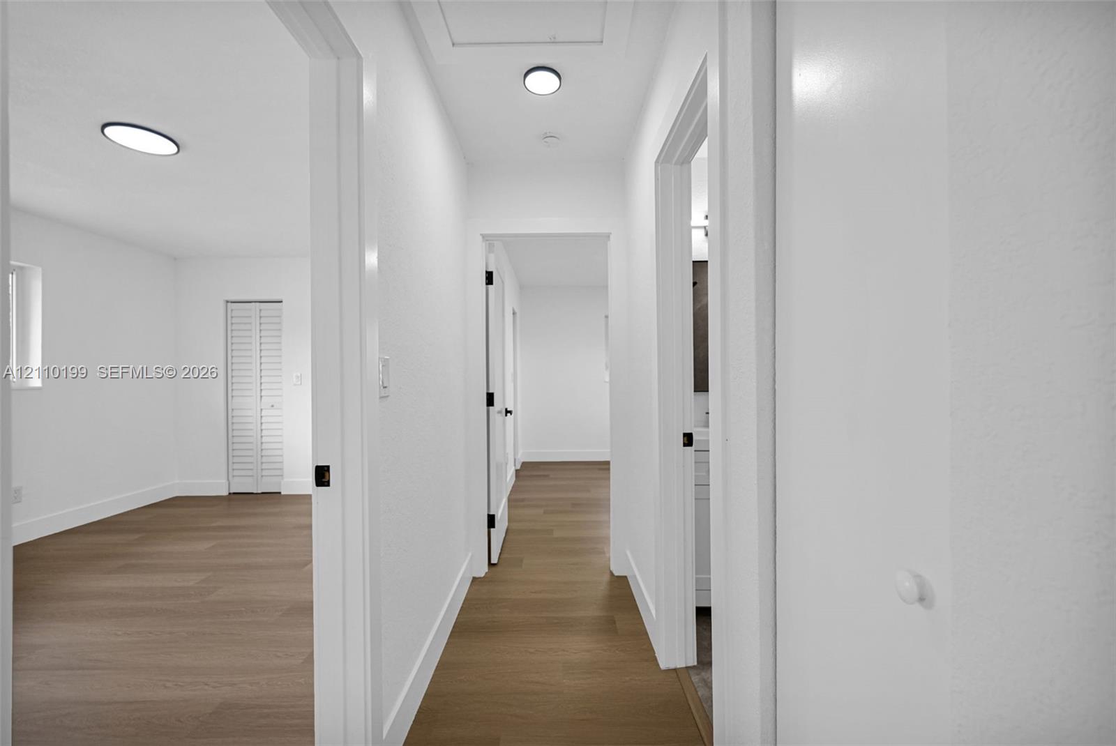 Hallway featuring wood-finish flooring, white walls, flush-mount circular lighting, and louvered closet doors