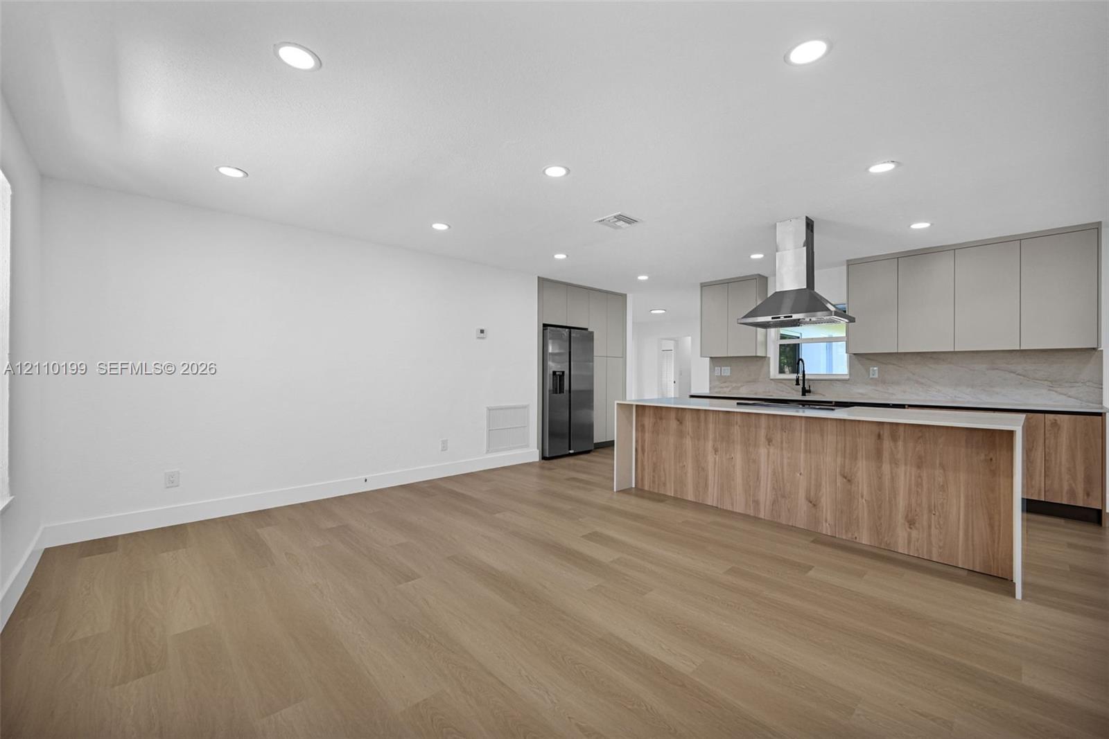 Kitchen featuring wood-finish cabinetry, an island with a light-toned countertop, a stainless steel chimney-style range hood, integrated refrigerator, and wood-finish flooring