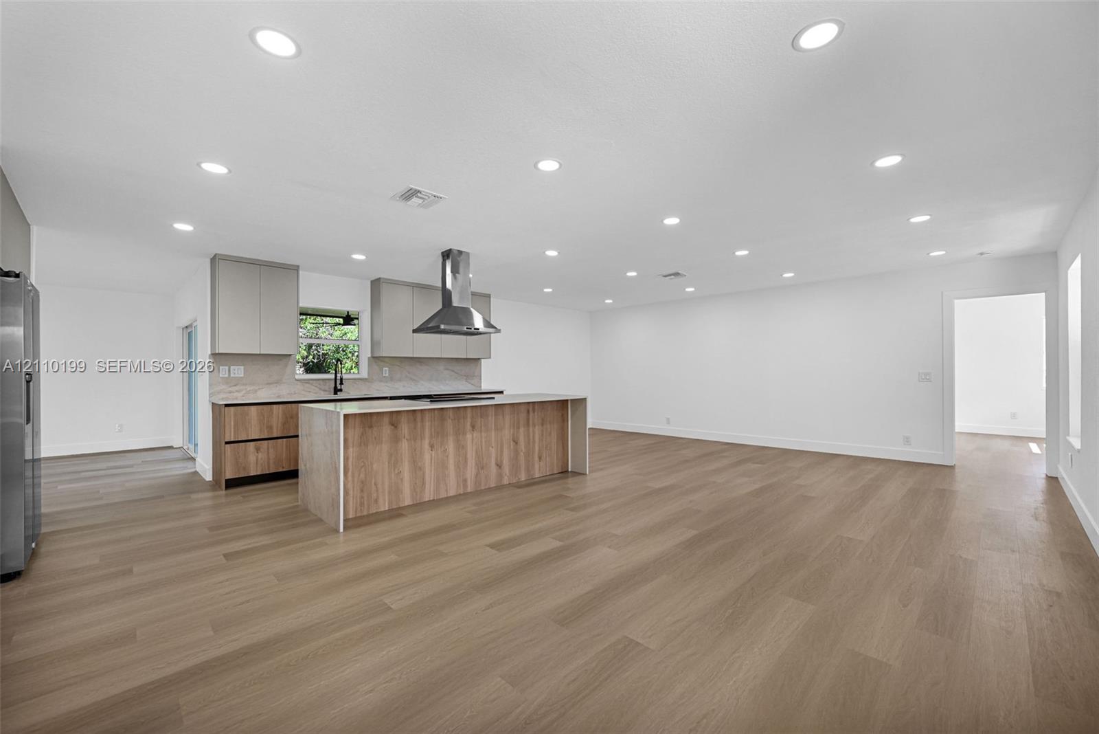 Kitchen featuring flat-panel cabinetry, a center island with waterfall-edge countertop, stainless steel range hood, stone-finish backsplash, and wood-finish flooring