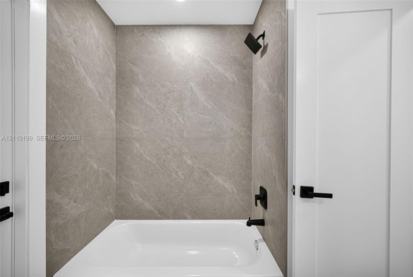 Full-height neutral stone-finish tile surround, white soaking tub, and matte black plumbing fixtures