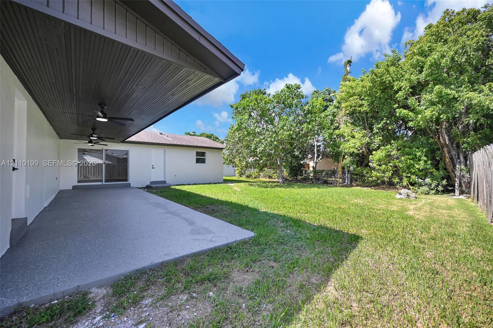 Covered concrete patio with beadboard ceiling and dual built-in ceiling fans, featuring a landscaped backyard with mature trees and perimeter wood fencing