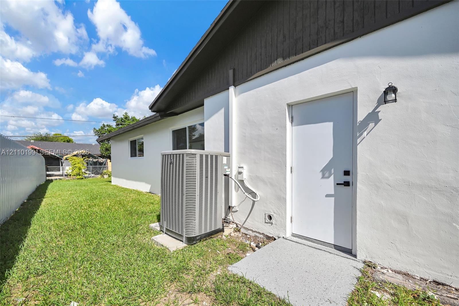 Exterior side yard featuring white stucco siding, dark wood-finish eaves, an exterior access door with matte black hardware, a wall-mounted sconce, and a central air conditioning unit
