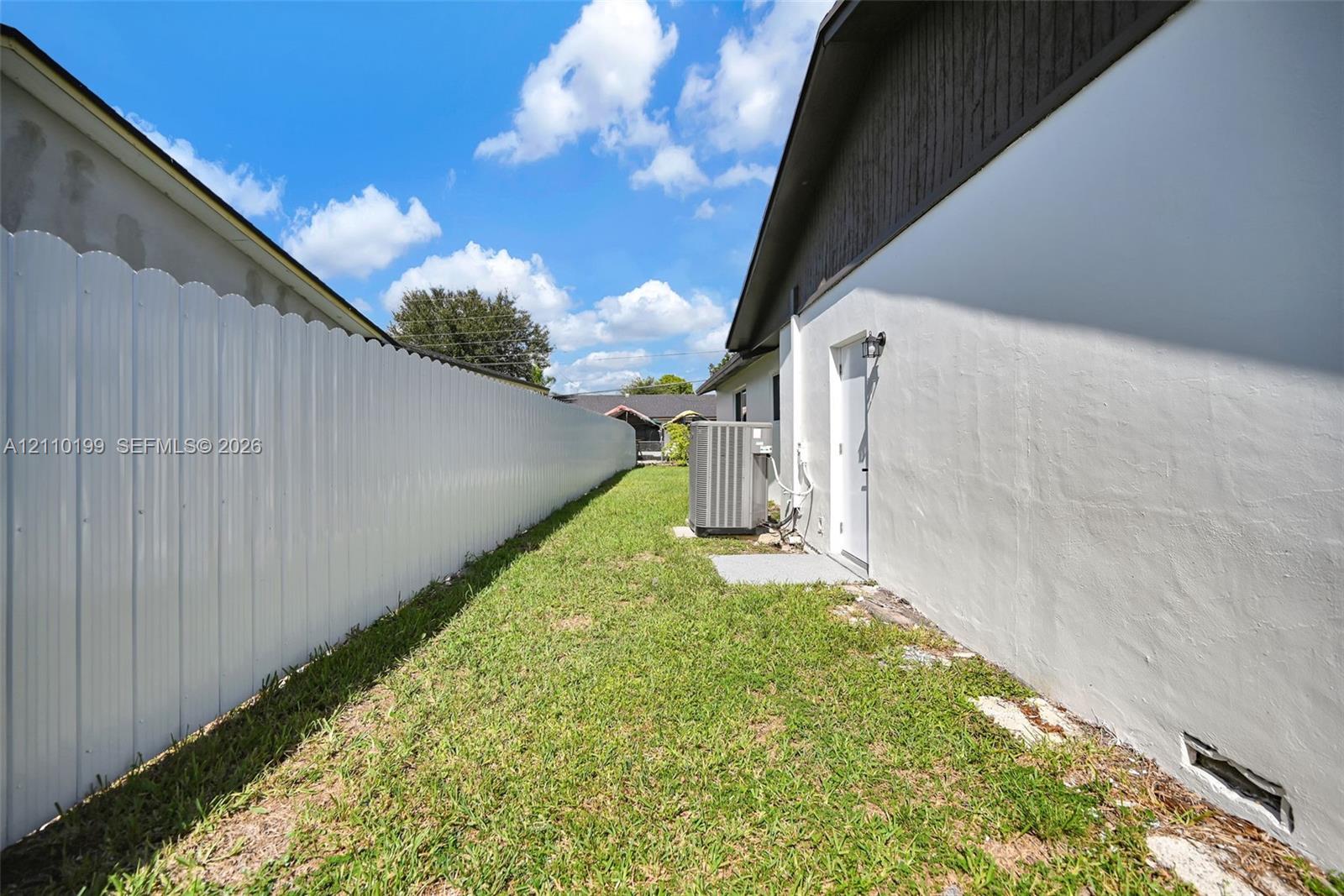 Side yard featuring a white corrugated metal fence, grass lawn, and exterior wall-mounted lighting