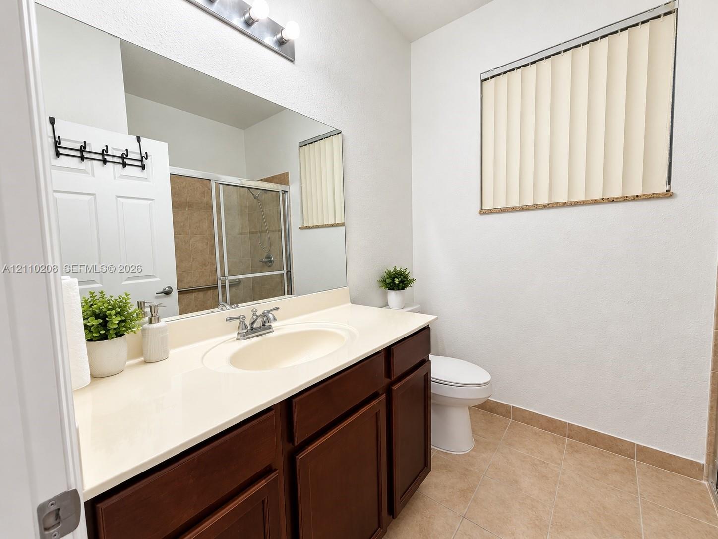 Vanity featuring dark wood cabinetry, an integrated sink with chrome faucet, and a large wall-mounted mirror with multi-bulb overhead lighting, complemented by tile flooring and a sliding glass shower enclosure