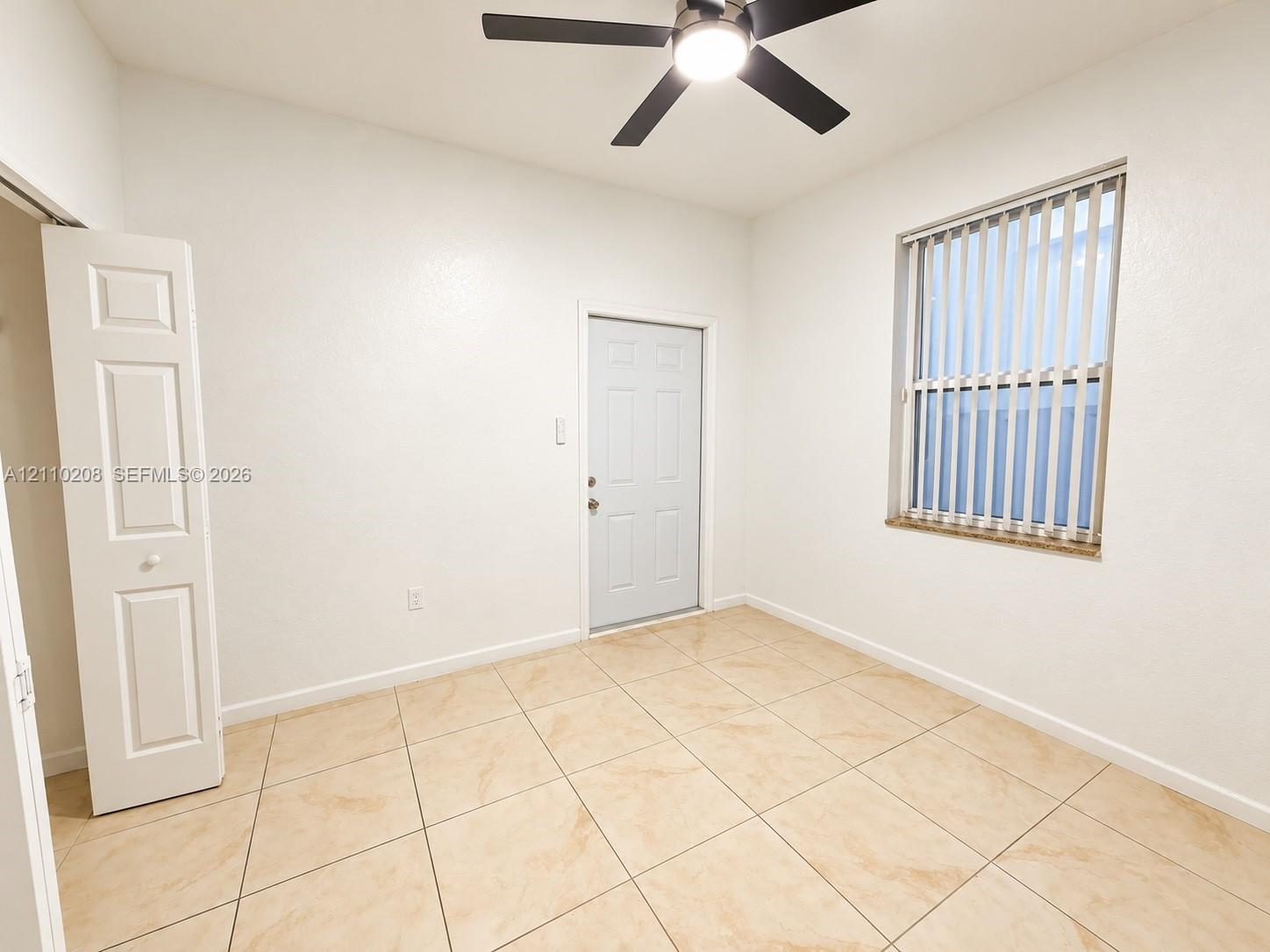 Room featuring large-format neutral tile flooring, ceiling fan with integrated lighting, window with vertical blinds, and a built-in bifold closet
