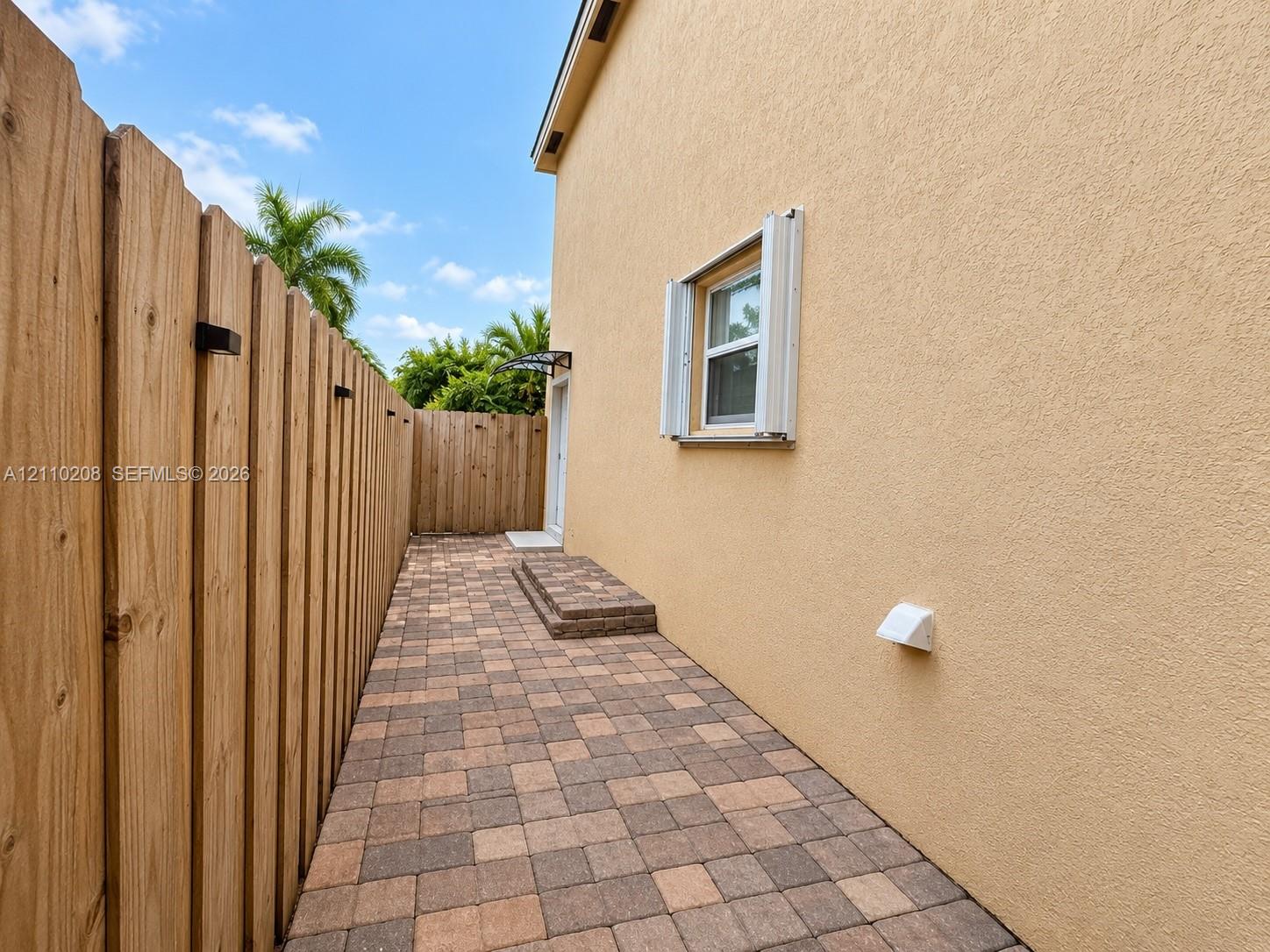 Stucco exterior wall with a single-pane window featuring accordion shutters, multi-toned concrete paver walkway, and full-height wood-slat privacy fencing
