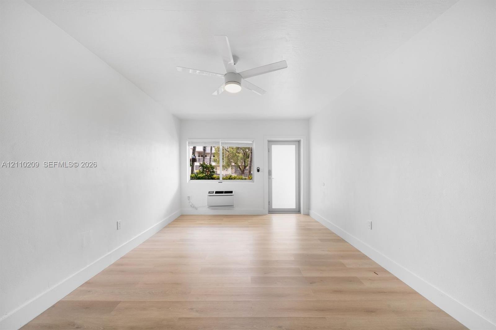 Bright interior featuring wood-finish flooring, a contemporary ceiling fan, a large window, and an integrated wall-mounted climate control unit