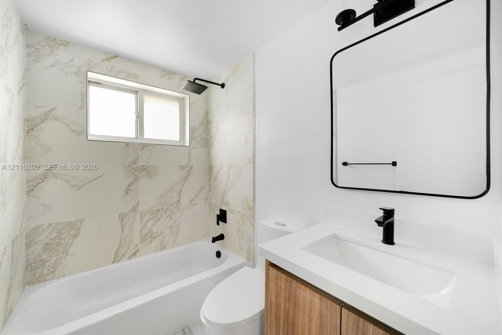 Bathroom featuring a wood-finish vanity, white countertop with integrated sink, matte black faucet, and shower-tub combination with light-toned patterned tile surround and matte black hardware