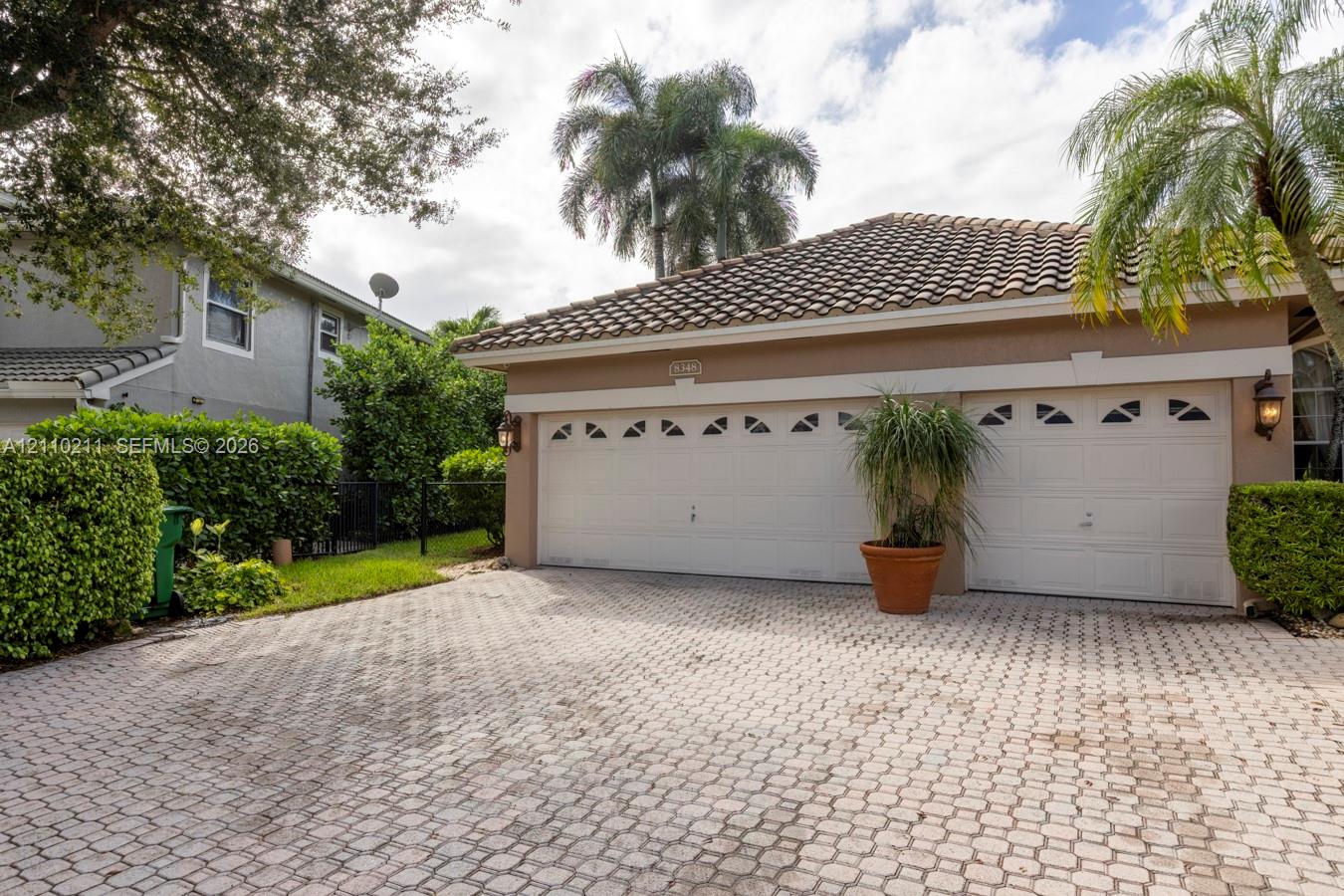 Paver driveway leading to a two-car garage with arched decorative windows, tile roof, and exterior wall-mounted carriage lights