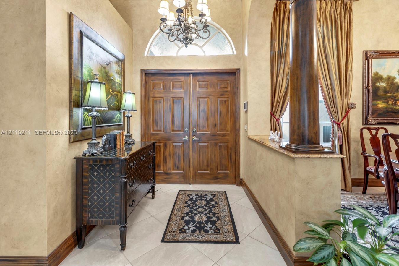 Foyer featuring double wood entry doors, an arched transom window, a decorative chandelier, tile flooring, and a stone-topped half wall with an ornamental column