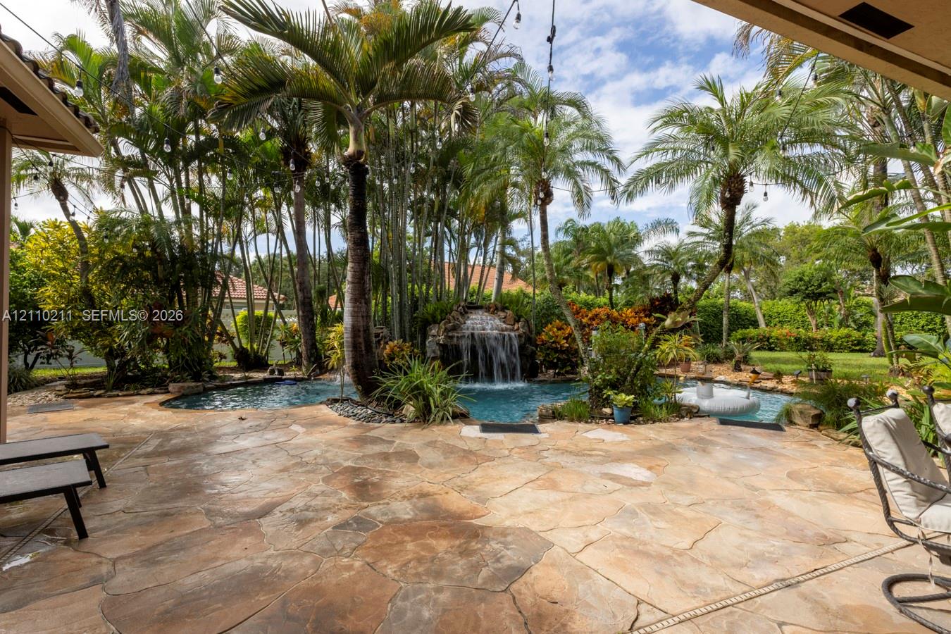 Flagstone patio area featuring a free-form pool with a rock waterfall feature, mature palm trees, and lush tropical landscaping