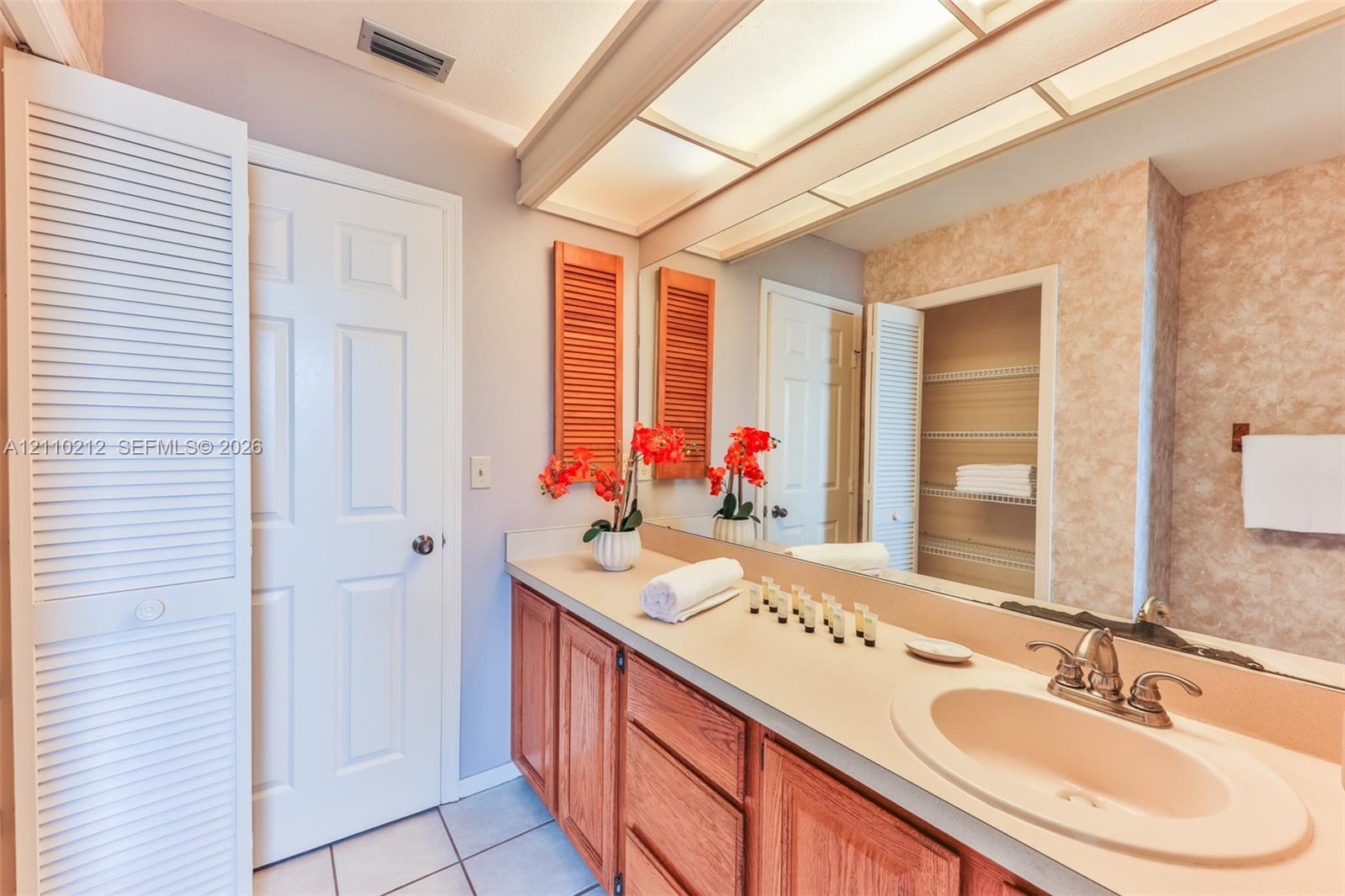 Bathroom vanity featuring wood cabinetry, a neutral countertop with an integrated basin, a full-width wall mirror, recessed overhead lighting, and built-in louvered-door cabinetry