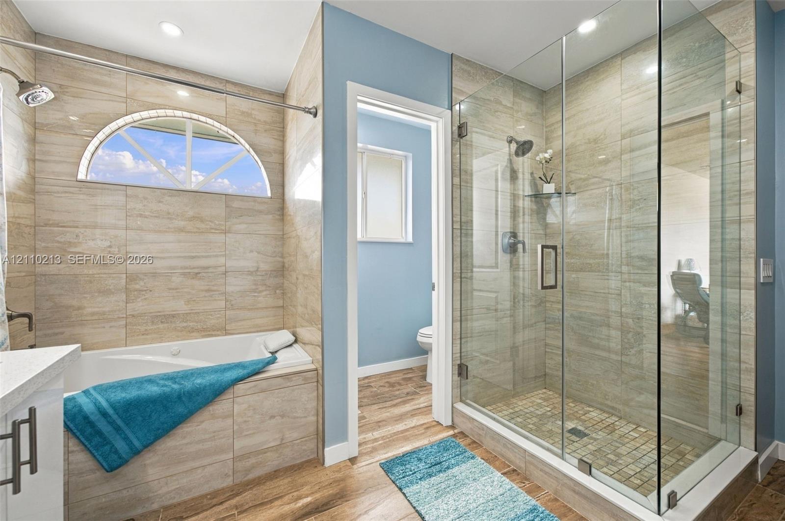 Walk-in shower with a glass enclosure, jetted soaking tub, arched window, and large-format tile surround