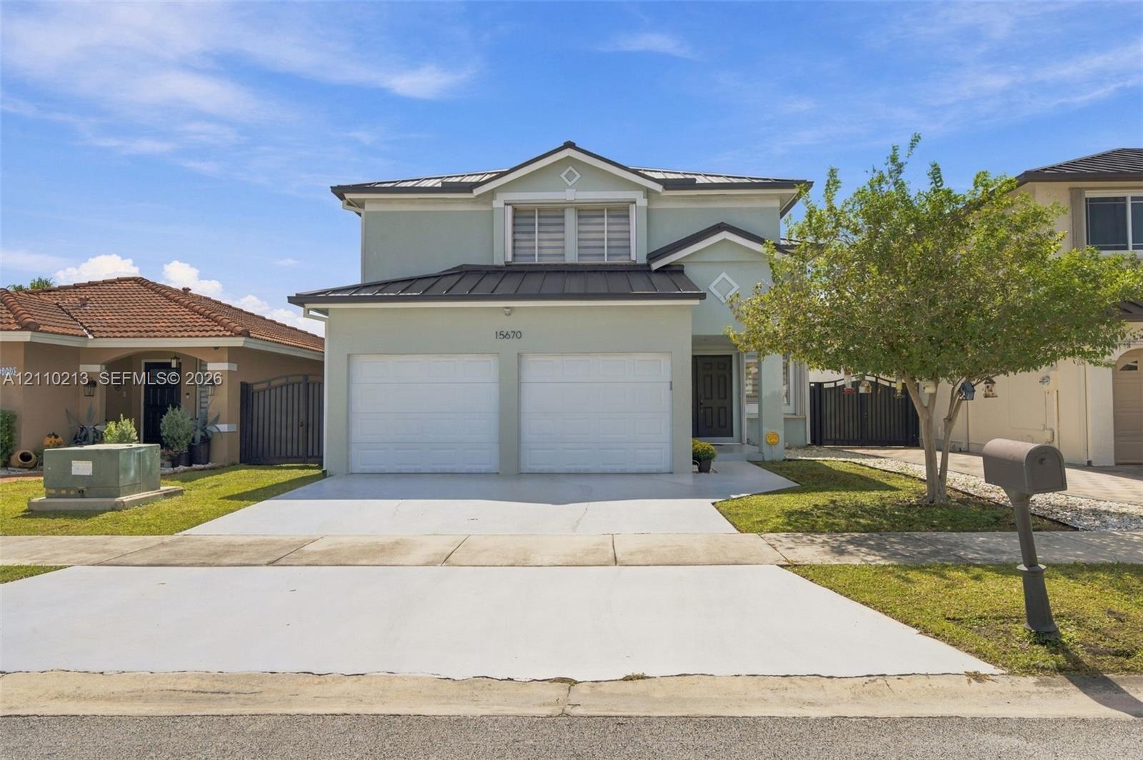 Two-story residence featuring a light-toned stucco exterior, a metal roof, an attached two-car garage, and a paved driveway Two-story residence featuring a light-toned stucco exterior, a metal roof, an attached two-car garage, and a paved driveway