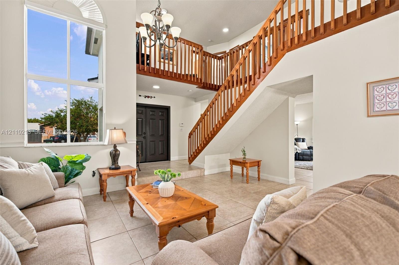 Foyer featuring a double-height ceiling, large multi-pane windows, wood-finish staircase railing, tiled flooring, and a multi-arm chandelier