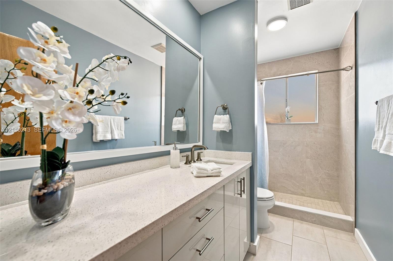 Bathroom featuring a white countertop vanity with under-mount sink, large wall-mounted mirror, polished metal hardware, and a walk-in shower with stone-finish tile surround