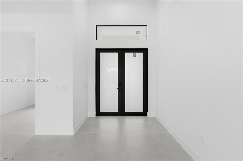 Double front doors featuring matte black frames with glass panel inserts, complemented by a rectangular transom window and large-format tile flooring