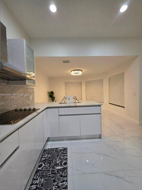 Kitchen featuring glossy white flat-panel cabinetry, light-toned stone-finish flooring, a textured stone-tile backsplash, and recessed overhead lighting