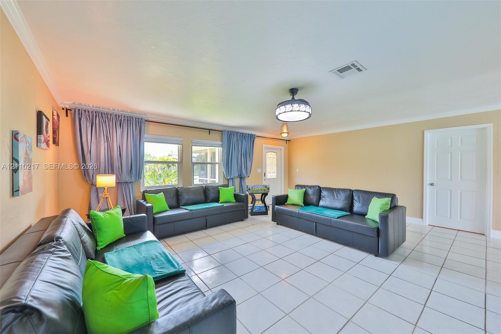 Living area featuring large format white tile flooring, crown molding, double windows with wall-mounted curtain hardware, and a ceiling fan with integrated lighting