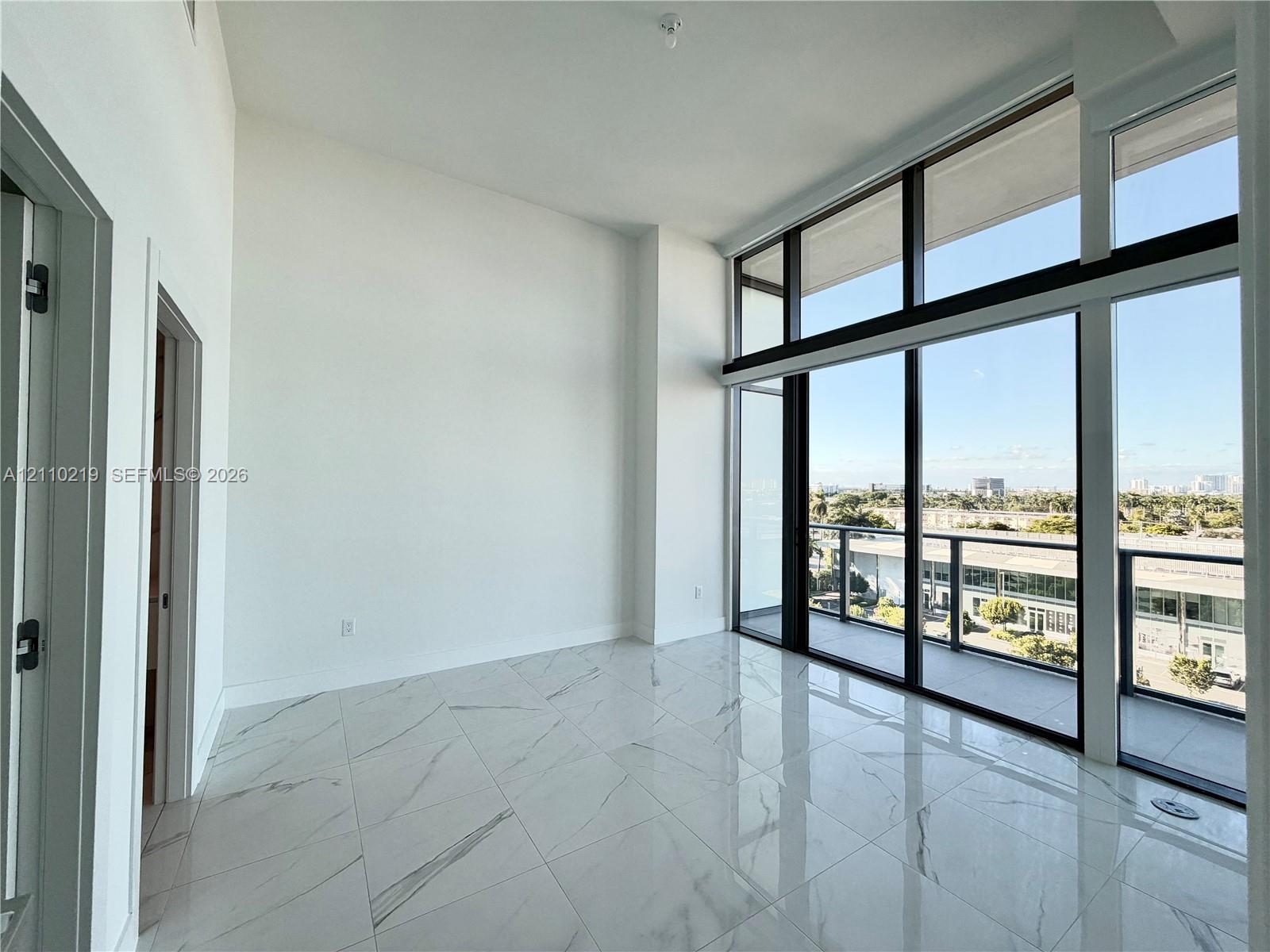 High-ceilinged room featuring large-format polished stone-finish floor tiles, floor-to-ceiling windows with dark framing, and a balcony