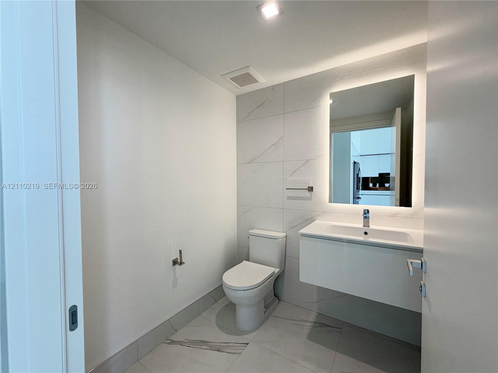 Floating vanity with an integrated sink, large-format tile flooring, wall-to-wall decorative tile backsplash, and LED-backlit mirror