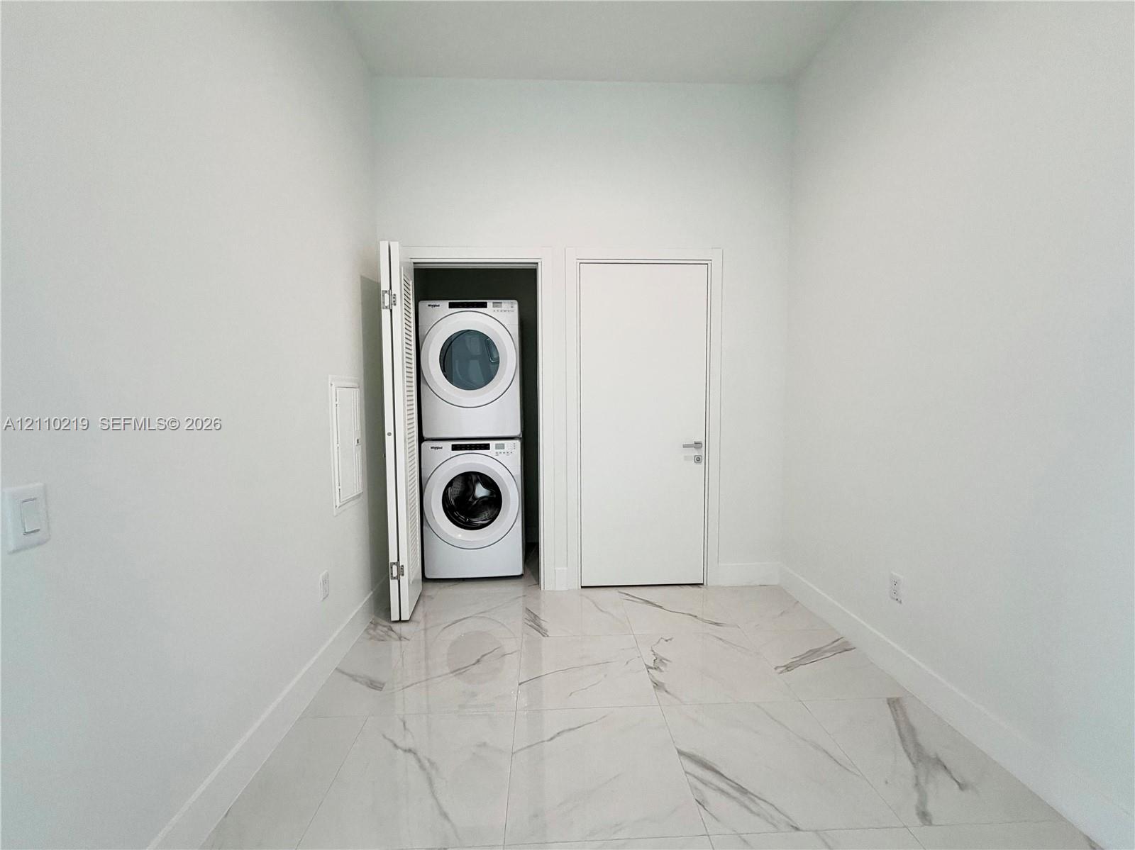 Utility area featuring large-format tile flooring with a neutral veined pattern, crisp white walls, and a recessed closet housing a stacked washer and dryer unit